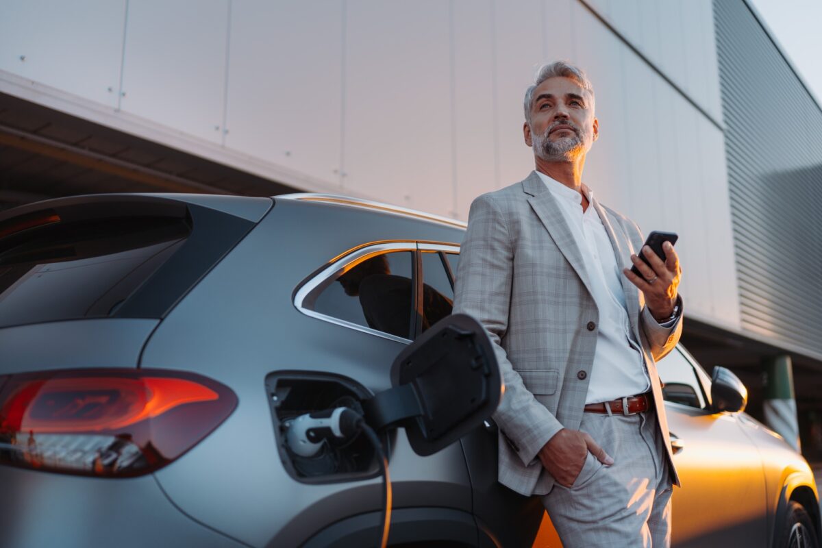 Homme d'affaires tenant un smartphone tout en chargeant sa voiture à une station de recharge de véhicules électriques, gros plan.
