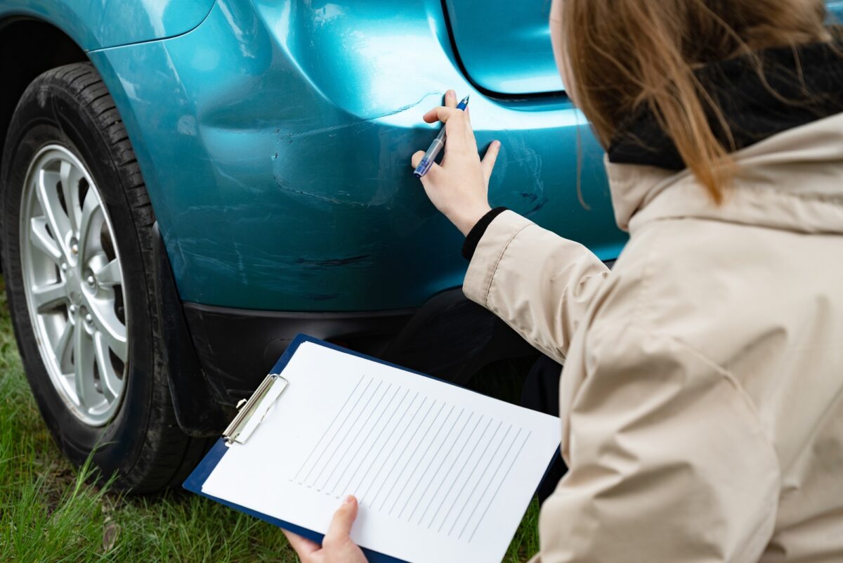 Personne inspectant les dommages causés au pare-chocs d'une voiture à l'aide d'un presse-papiers, à l'extérieur.