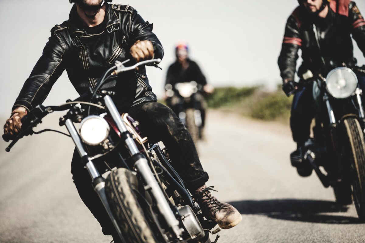 Trois hommes portant des blousons de cuir et conduisant des motos de type "cafe racer" sur une route de campagne.